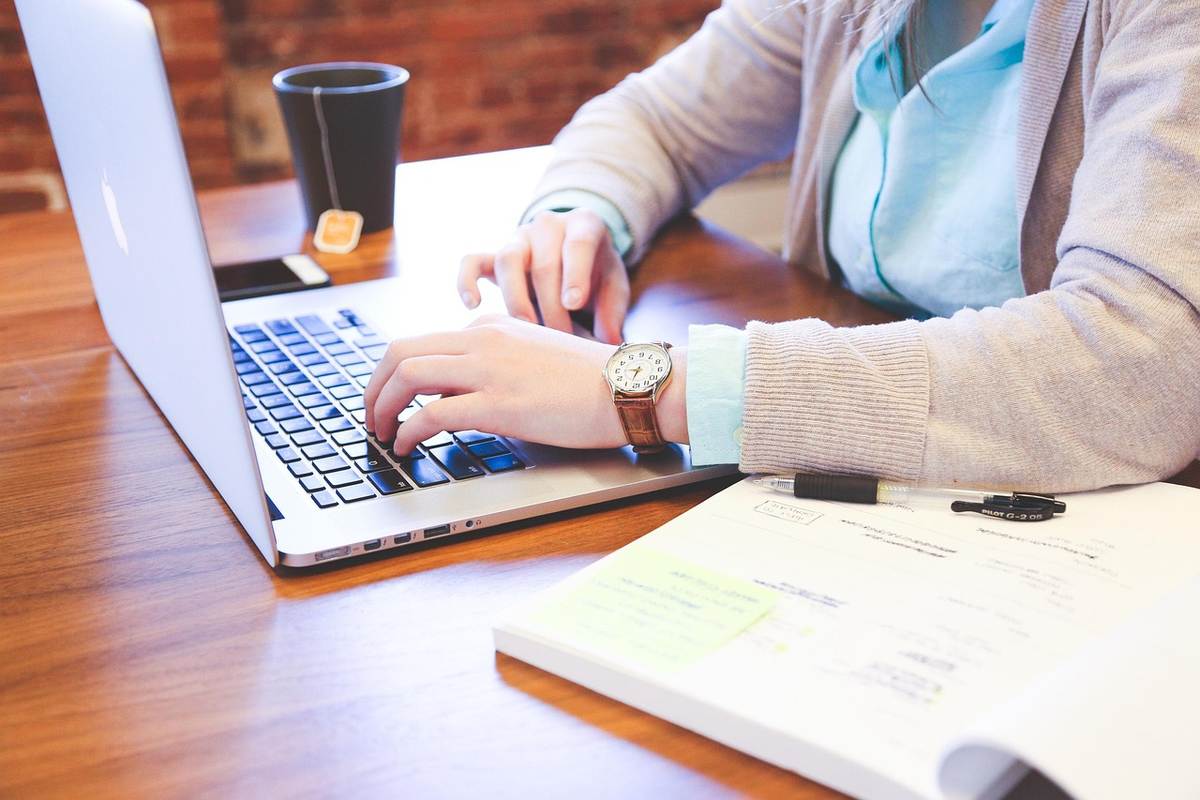 Une personne tape sur le clavier d'un ordinateur portable posé sur un bureau en bois, avec un verre à côté