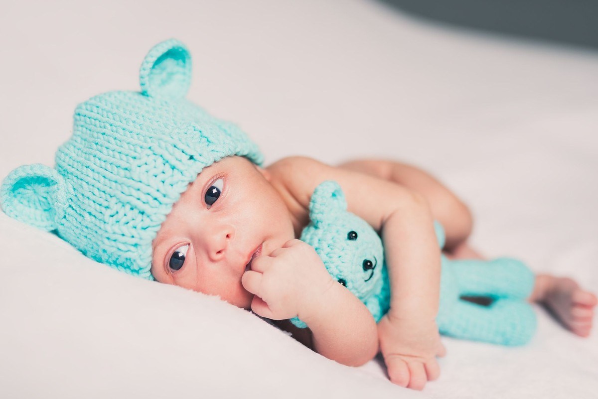 Un nourrisson avec bonnet d'ours et peluche bleue sur drap blanc