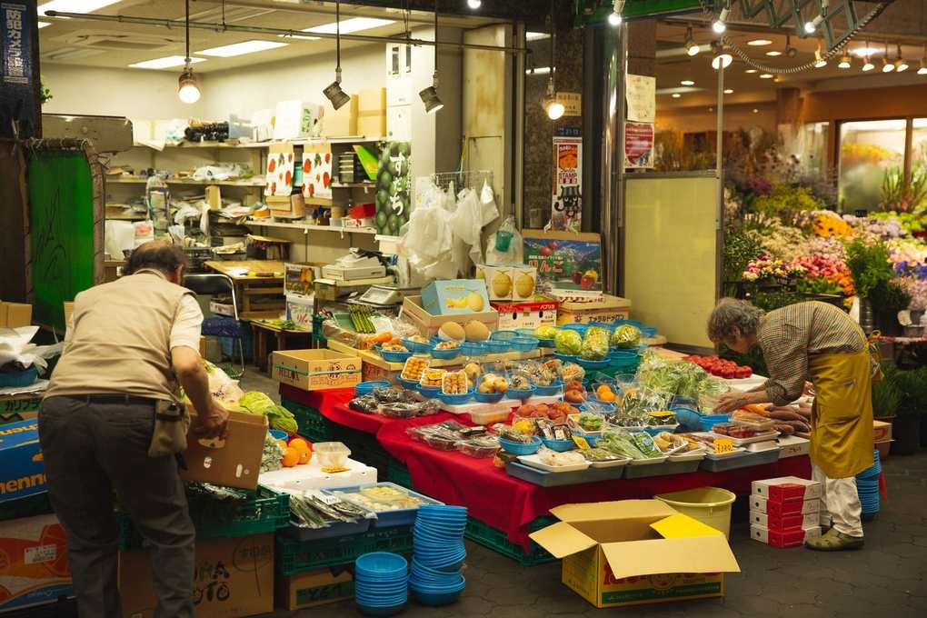 Marché avec étals de fruits et légumes, deux personnes en train de travailler