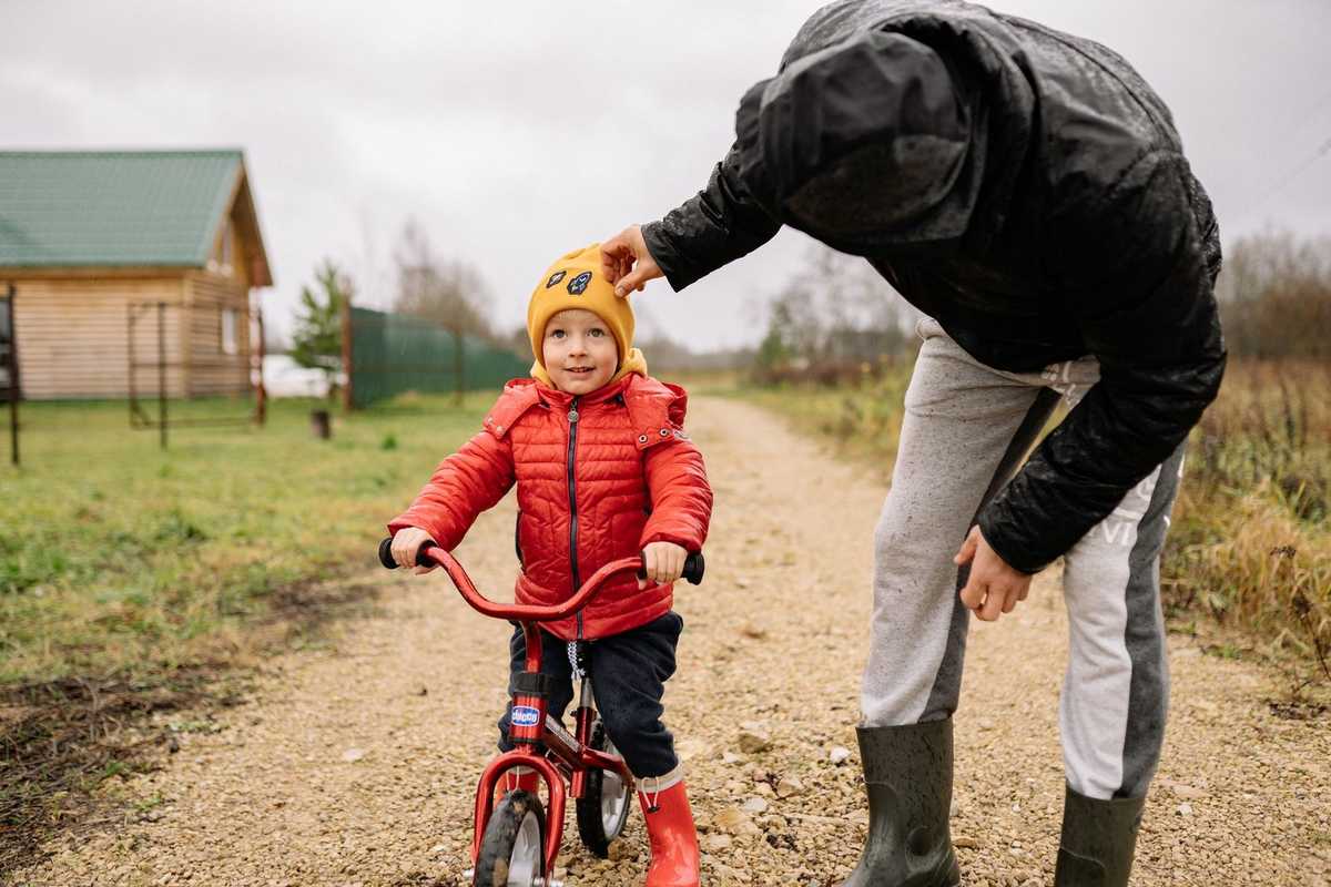 Enfant en rouge sur vélo miniature, adulte en noir ajuste son chapeau jauni, chemin de terre, maison verte Enfant en rouge sur vélo miniature, adulte en noir ajuste son chapeau jauni, chemin de terre, maison verte