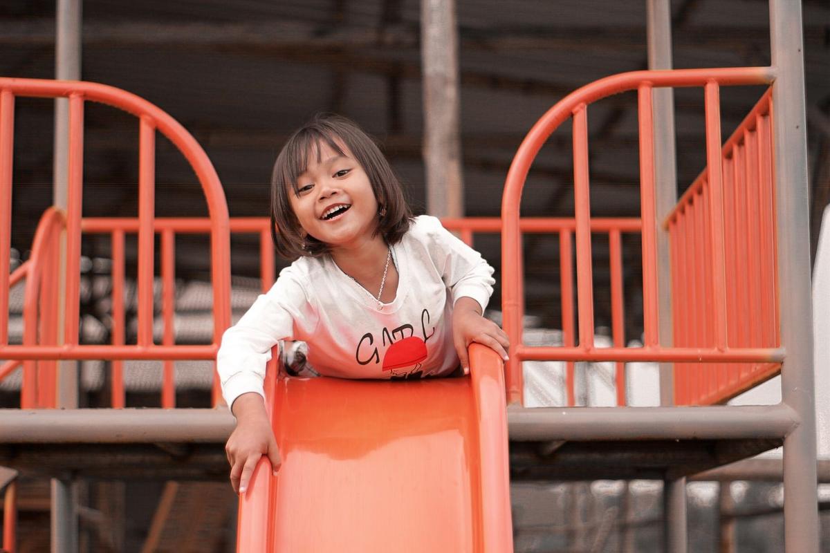 Enfant sur un toboggan orange, souriant, vêtu de blanc, cheveux courts Enfant sur un toboggan orange, souriant, vêtu de blanc, cheveux courts