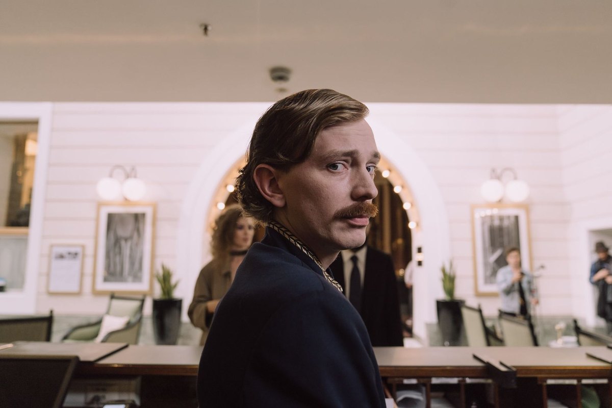 Homme en costume avec moustache dans un salon avec tables et portraits